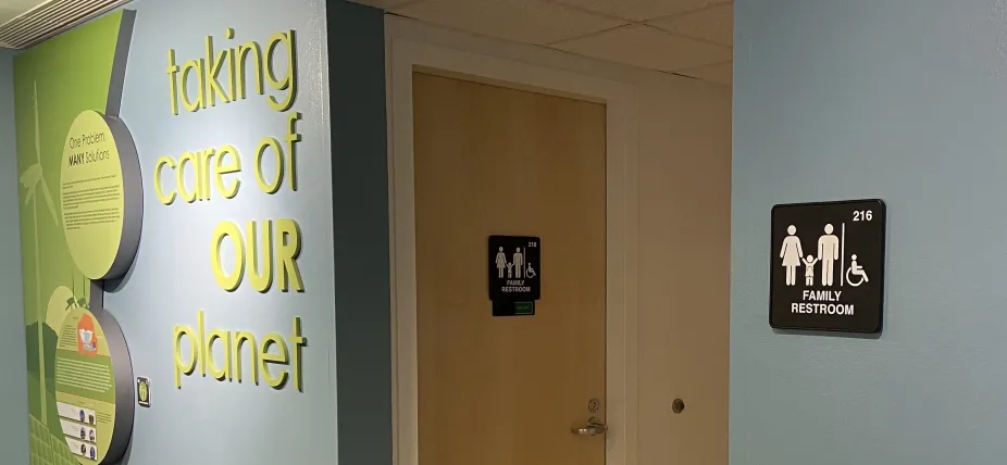 A photo of the accessible restroom door which shows a sign on it that says “Family Restroom.” This bathroom is right next to an exhibit wall that reads “taking care of our planet.”