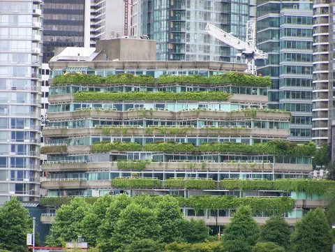 This is an image of a multi-storied apartment building surrounded by tall buildings in a city. The terraces of the apartment building are covered in vegetation, and there are lots of trees around the perimeter of the building as well.